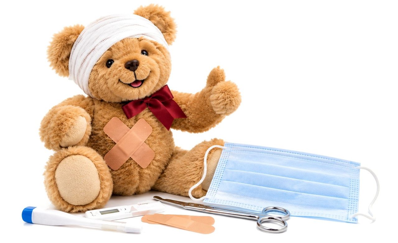 Teddybär mit Kopfverband und Pflaster, daneben Einmal-Maske und Thermometer